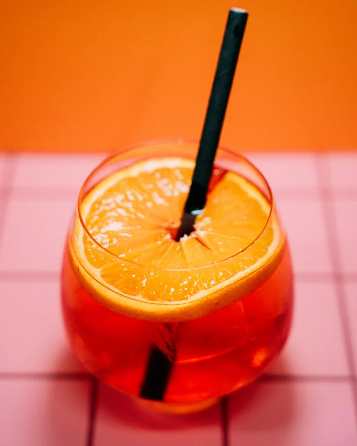 Glitter Spritz – schimmernder Orangen-Spritz mit Orangenscheibe auf rosa Fliesen, Signature Drink der Adieu Bar Prenzlauer Berg