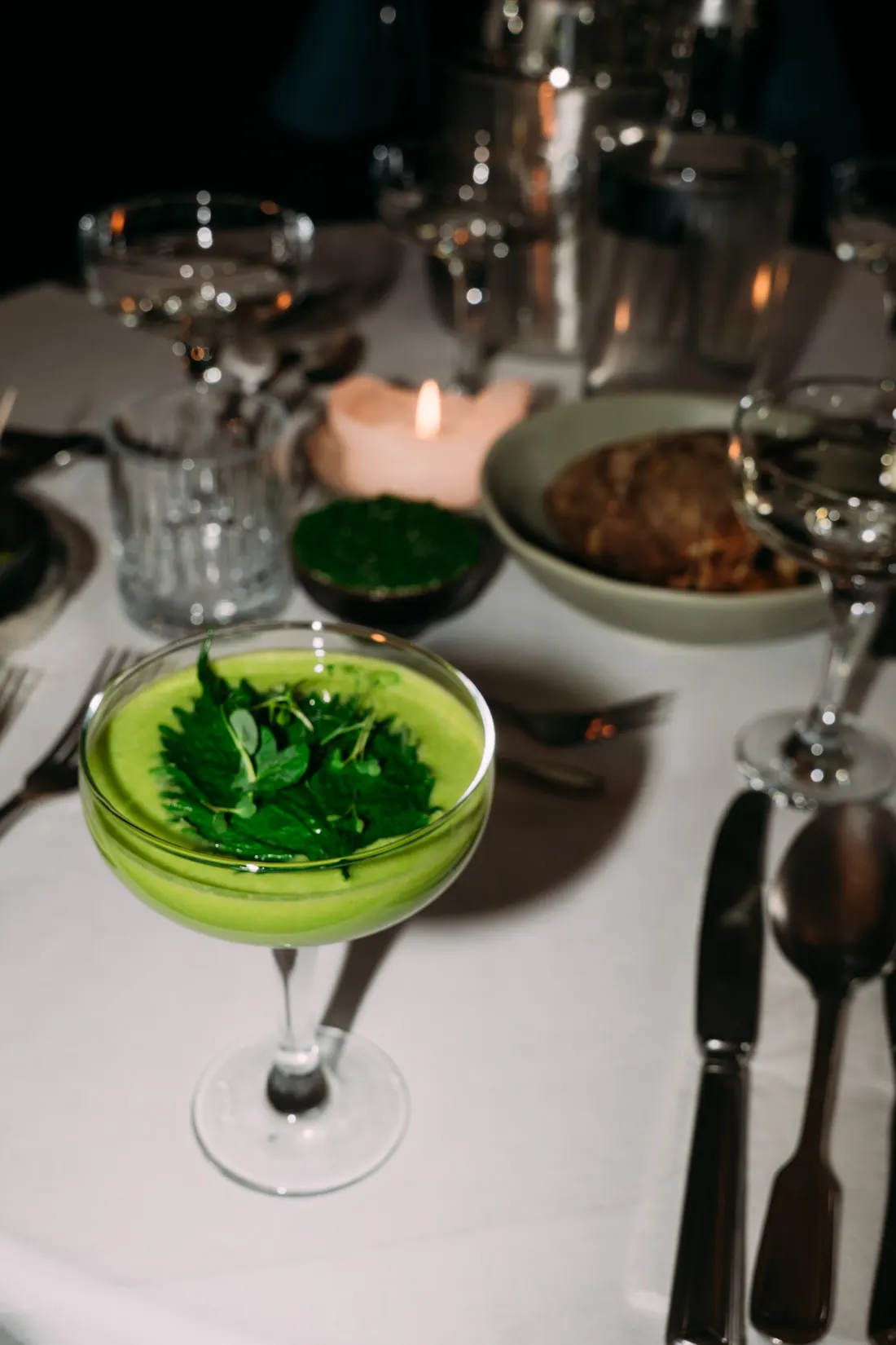 Kalte grüne Suppe in einer Coupette, mit Kräutern bedeckt, auf weißem Tischtuch bei Kerzenlicht